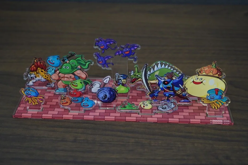 ドラゴンクエストシリーズの様々なモンスターやキャラクターのアクリルスタンドが、レンガ調の台座に並べられています。スライム、ゴーレム、キングスライムなど、お馴染みのキャラクターたちが集結した賑やかなゲームグッズです。