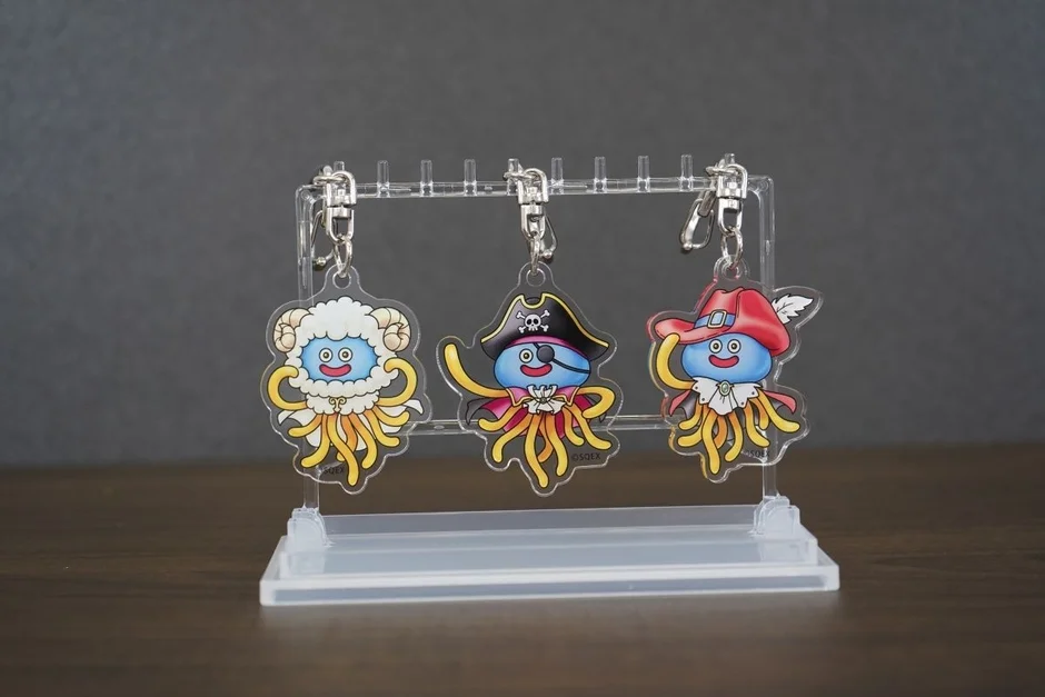ドラゴンクエストシリーズのキャラクター「タコメット」をモチーフにしたアクリルキーホルダー3点。羊、海賊、冒険家風の衣装をまとっており、透明なディスプレイスタンドに飾られている可愛らしいゲームグッズです。