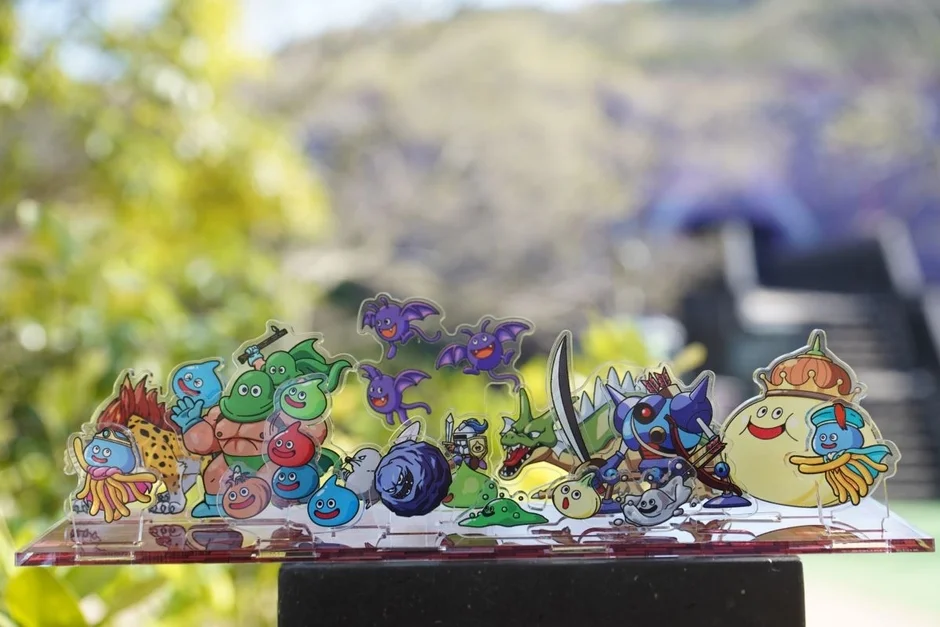 ドラゴンクエストシリーズの様々なモンスターたちがデザインされたアクリルスタンドが、自然豊かな屋外で撮影されています。スライムやゴーレム、ドラキーなど、多くの人気キャラクターが集まっています。